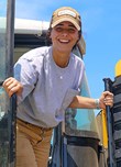Une ouvrière agricole descend d'un tracteur jaune en souriant.